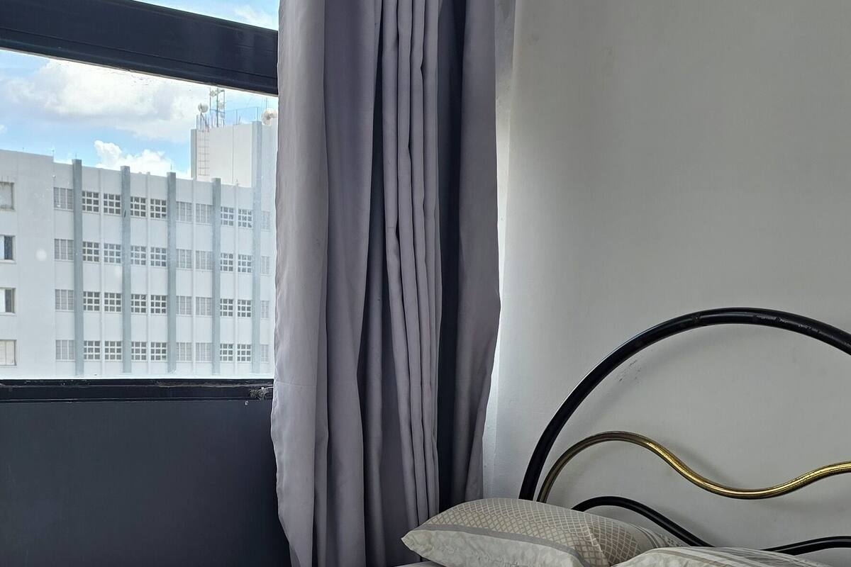 Una cama bien hecha con colcha y almohadas blancas, cabecero de metal negro y vistas a un edificio a través de una ventana con cortinas.