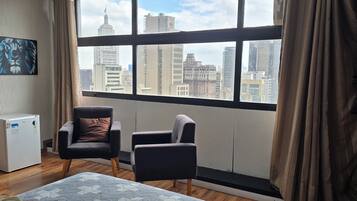 A room with a large window, two chairs, a small white appliance, and a painting on the wall.