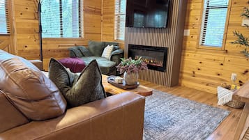 Cozy cabin living room with fireplace, TV, and bright woodland views