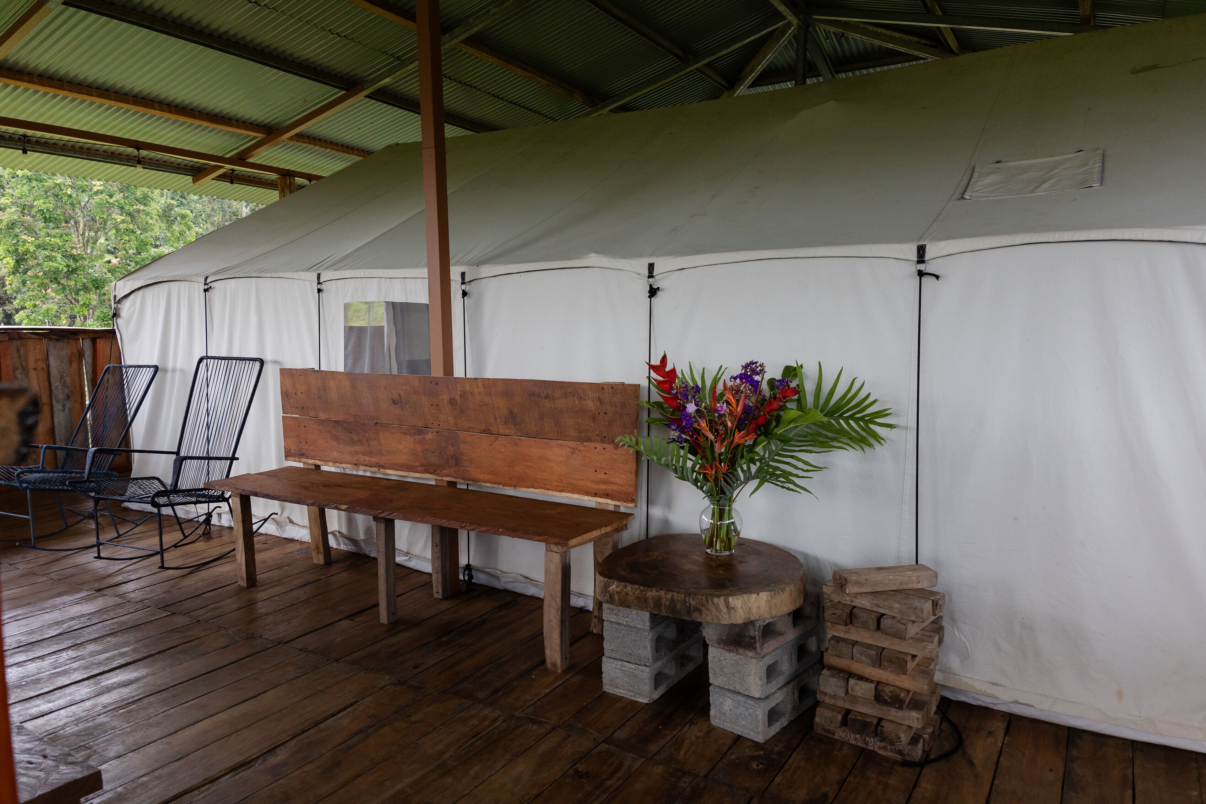 A wooden bench, a small table with a vase of flowers, and a stack of bricks on a wooden deck under a white tent.