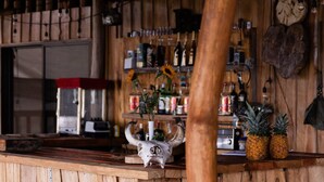 Un coin bar rustique en bois, avec un tabouret en bois, un grand tronc d’arbre au premier plan, et une étagère contenant diverses bouteilles et une horloge au mur.