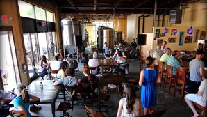 Un'ampia sala ristorante con soffitti alti e arredamento in stile industriale, caratterizzata da ampie finestre e una zona bar centrale.