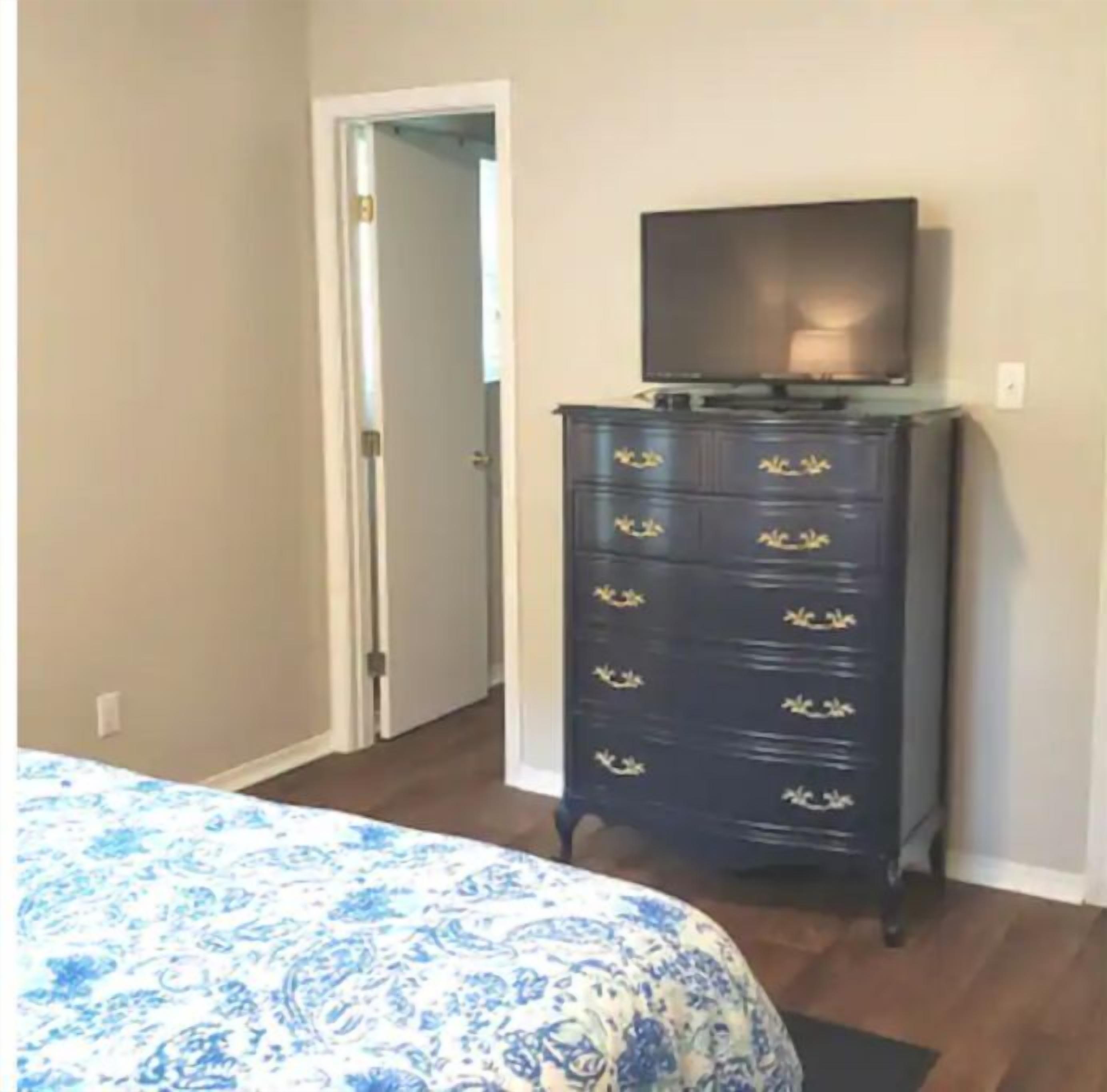 A bedroom with a bed, a dresser with drawers, a television on top, and a door leading to another room.