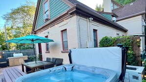A hot tub on a wooden deck, a barbecue grill, and a house with a green roof.