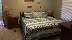 Un dormitorio con cama, dos mesitas de noche, un ventilador de techo, decoración de pared con brújula y una ventana con persianas.
