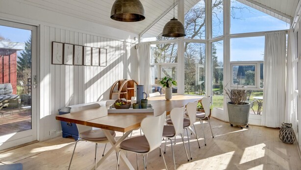 Ein heller Essbereich mit viel Sonnenlicht, einem Holztisch, weißen Stühlen, großen Fenstern und hängenden Pendelleuchten.