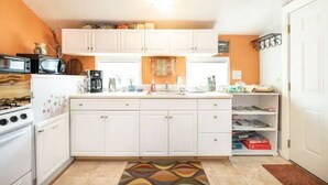 Cocina compacta con armarios blancos, estufa y microondas. Hay una ventana sobre el fregadero y una estantería con libros y objetos decorativos.