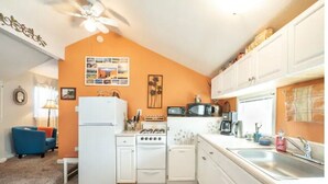 Cocina compacta con electrodomésticos blancos, microondas y un lavabo. En la zona de estar contigua hay una silla azul y una pequeña mesa.