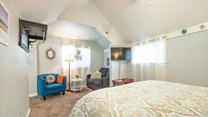 Un dormitorio con una cama grande, un sillón azul, un sillón gris, una mesita redonda y un televisor fijado en la pared.