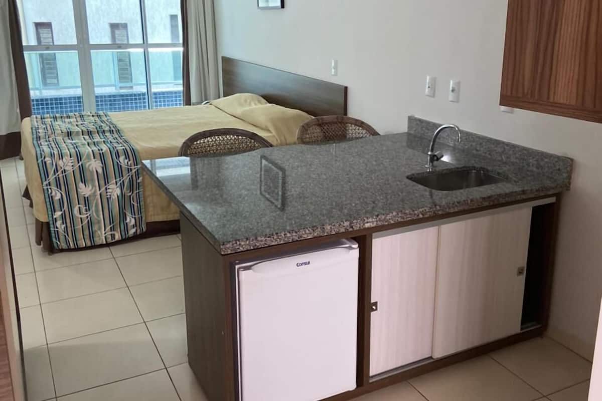 Une chambre d’hôtel compacte comprenant un coin cuisine, un lit avec une couvre-lit à rayures, une fenêtre avec des rideaux et une vue sur un bâtiment à l’extérieur.