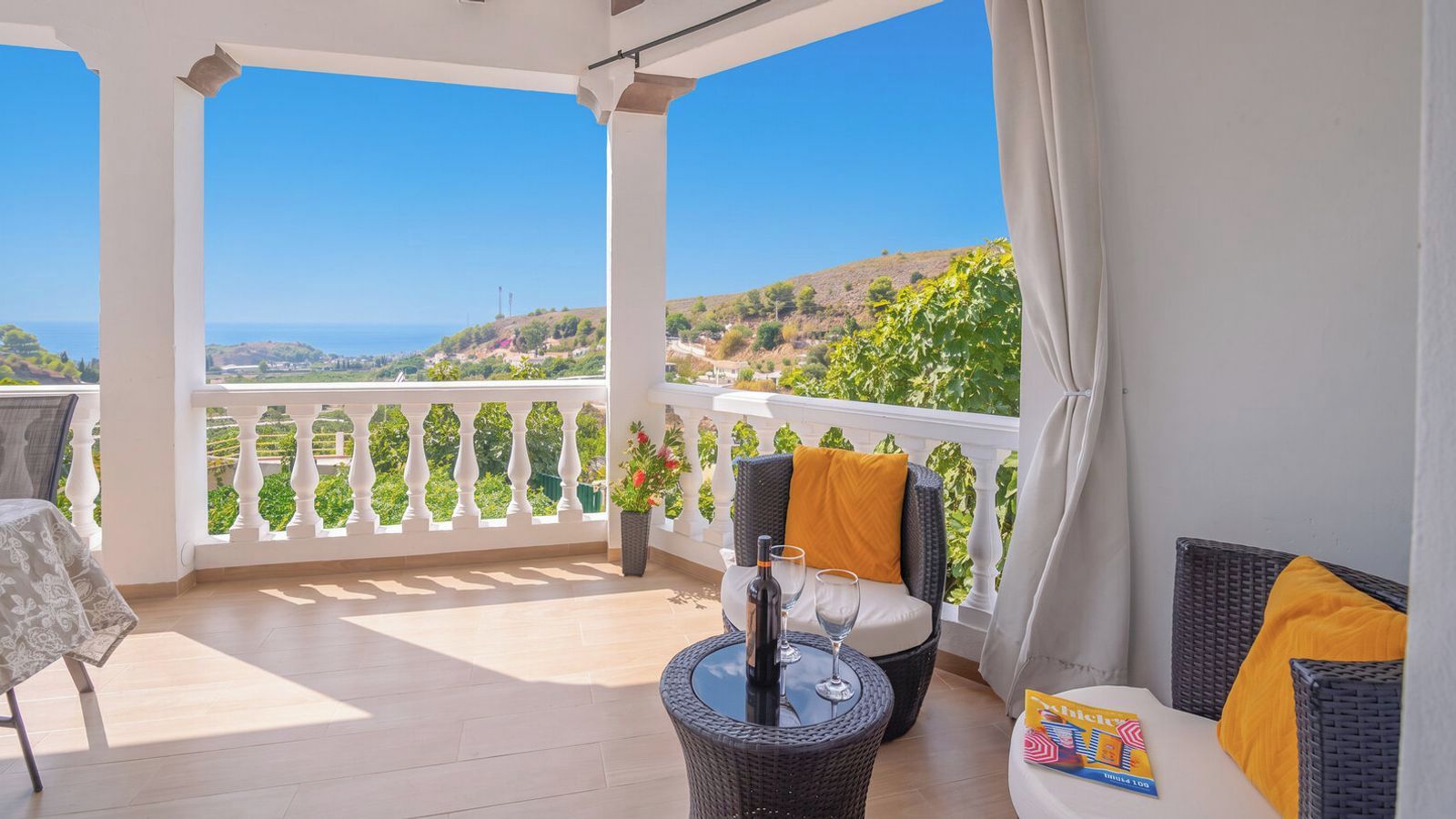 Un balcon avec une balustrade blanche, des meubles en osier et une table sur laquelle se trouvent une bouteille et des verres.