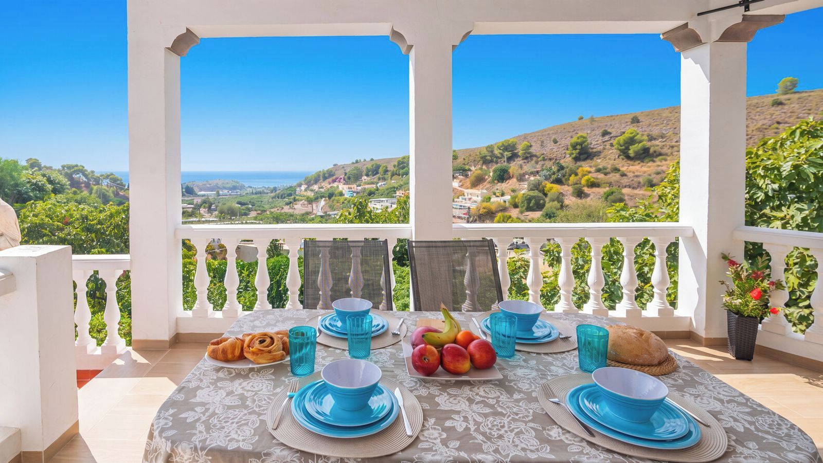 A table set for two with blue plates and glasses, a plate of fruit, and a croissant, overlooking a scenic landscape.