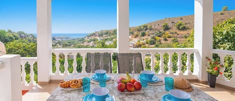A table set for two with blue plates and glasses, a plate of fruit, and a croissant, overlooking a scenic landscape.