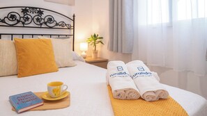 En pænt redt seng med en gul pude, et sammenrullet håndklæde med et logo, en bog og en kop på et bakke.