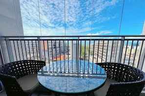 A balcony with a round glass table and two chairs, overlooking a cityscape.