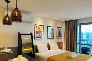 A hotel room with a bed, bedside table, and two hanging pendant lights. There are two framed artworks on the wall, a window with curtains, and a view of buildings outside.