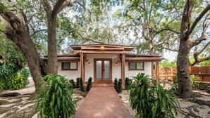 A two-story house with a wooden deck leading to the entrance, surrounded by large trees and potted plants. - Find Peace - Tiny House in Japanese Garden (Homestead)