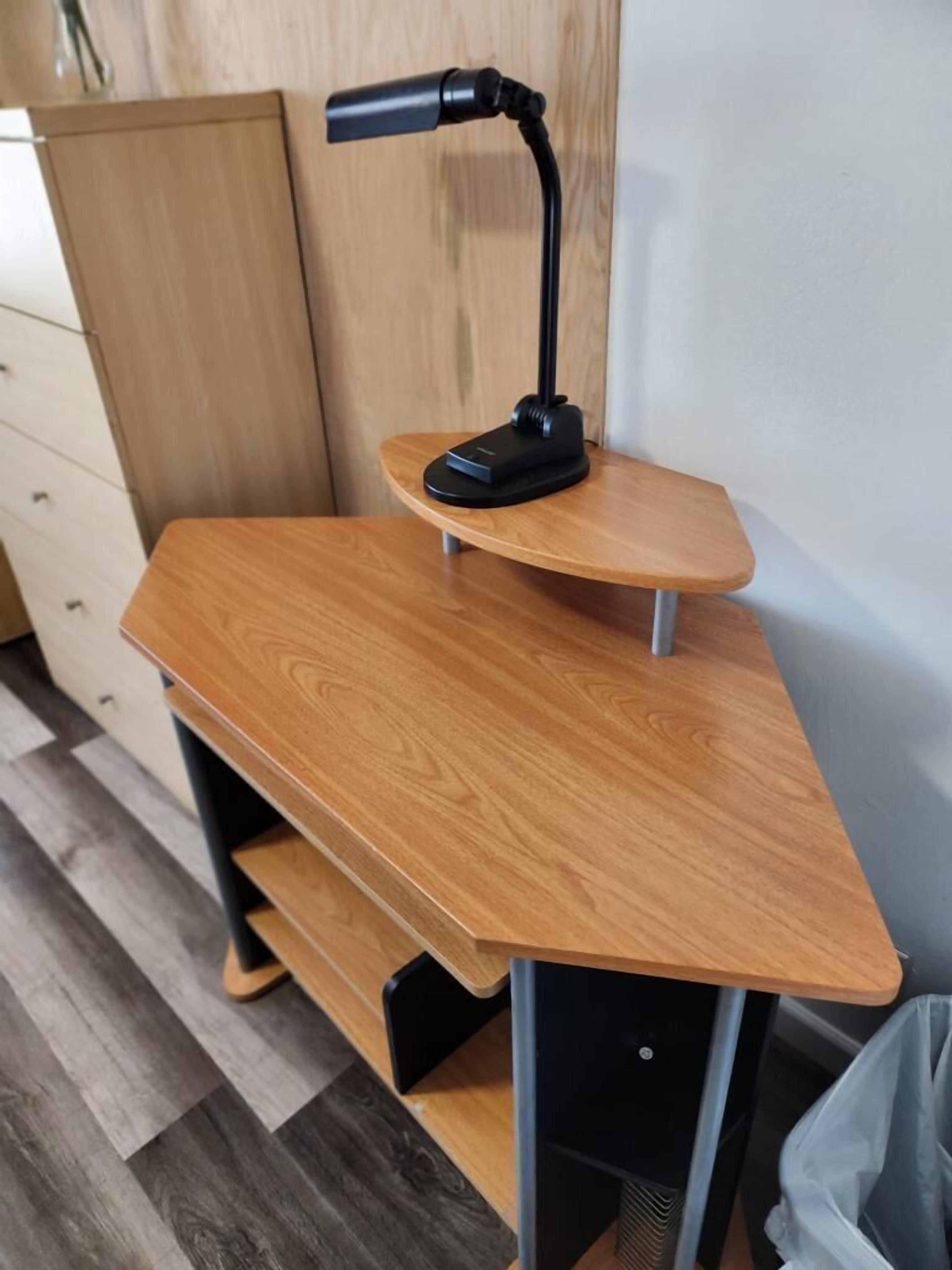 Ein Schreibtisch im Eckbereich mit einer schwarzen Lampe, einer Hollausziehvorrichtung und einem grauen Mülleimer.