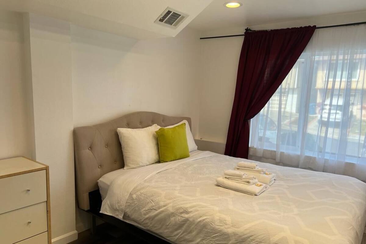 A neatly made bed with white bedding and a green pillow, a bedside table with drawers, and a window with sheer curtains.