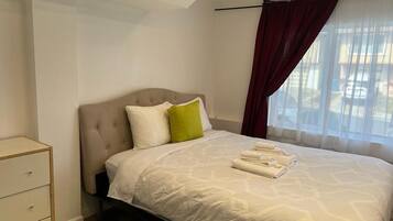 A neatly made bed with white bedding and a green pillow, a bedside table with drawers, and a window with sheer curtains.