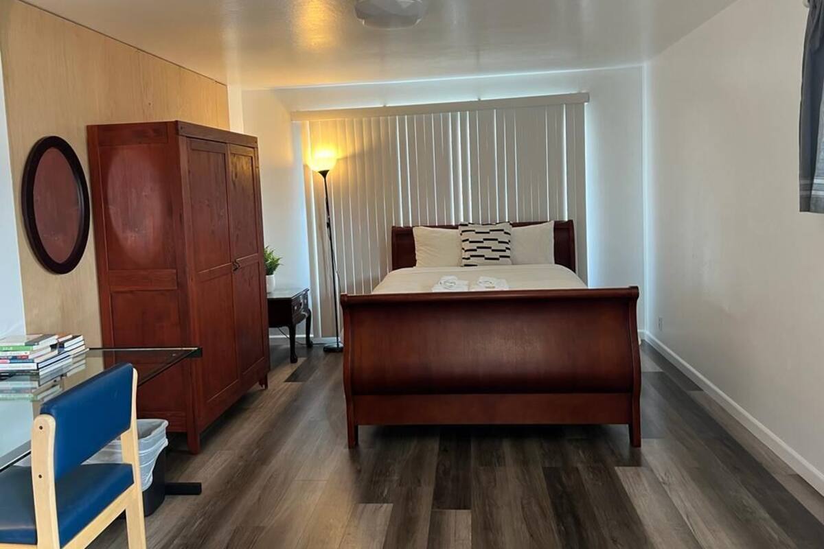 A bedroom with a wooden bed, a desk with books, a wardrobe, and a window with blinds.