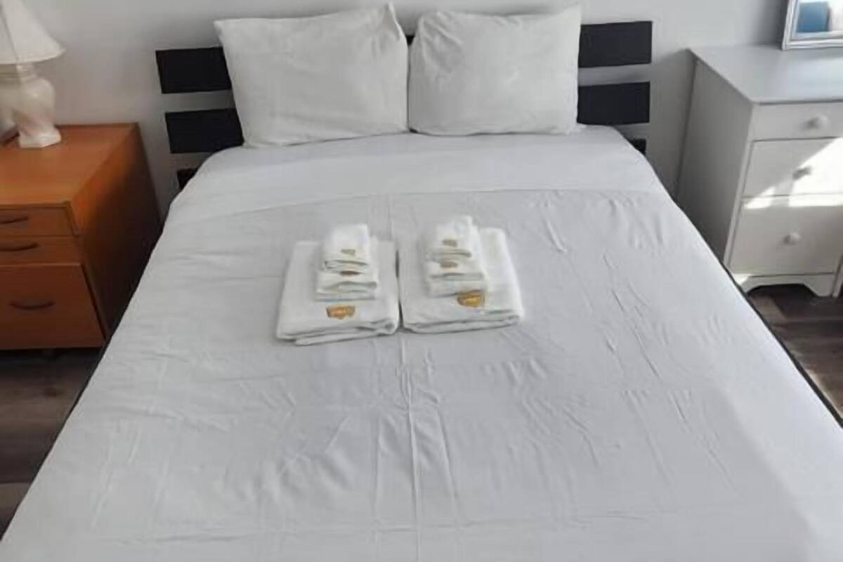 A neatly made bed with white linens and towels, a wooden nightstand with a lamp, and a white dresser with a mirror.