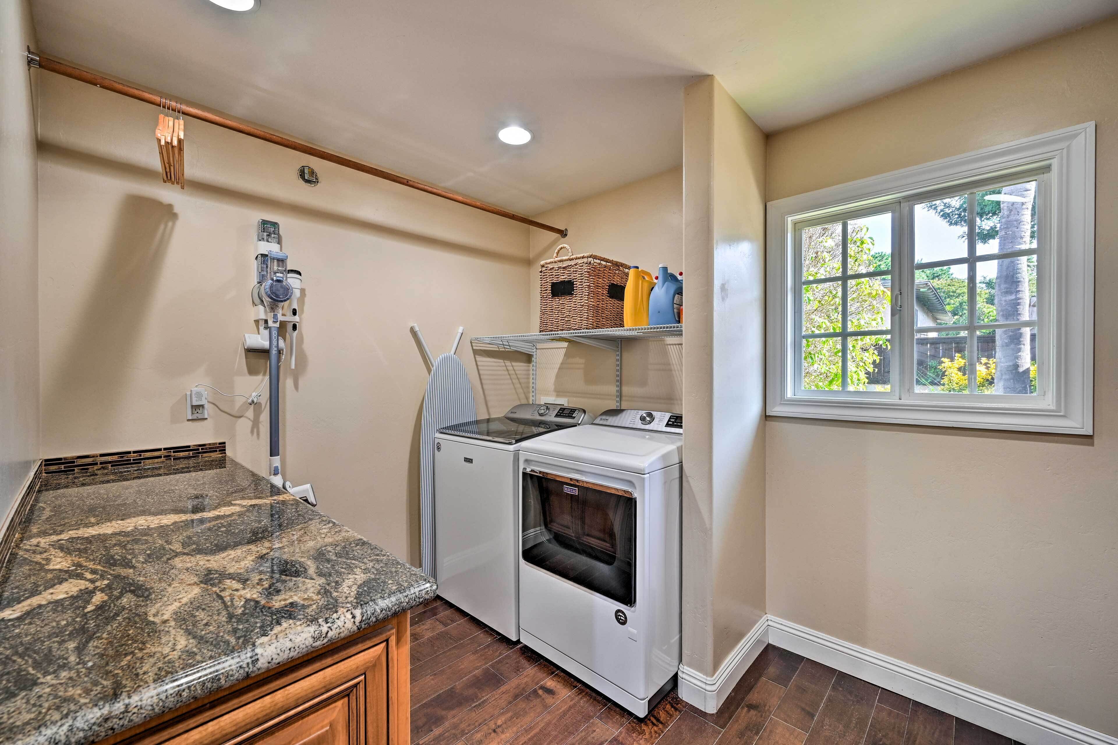 A laundry room with a granite countertop, two washing machines, a dryer, a window with a view, and a shelf with laundry supplies.