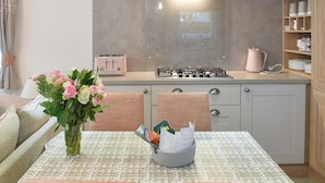 Een keuken met een gasfornuis, een broodrooster en een waterkoker op het aanrecht. Er staat een tafel met een bloemenvaas en een mand met post.