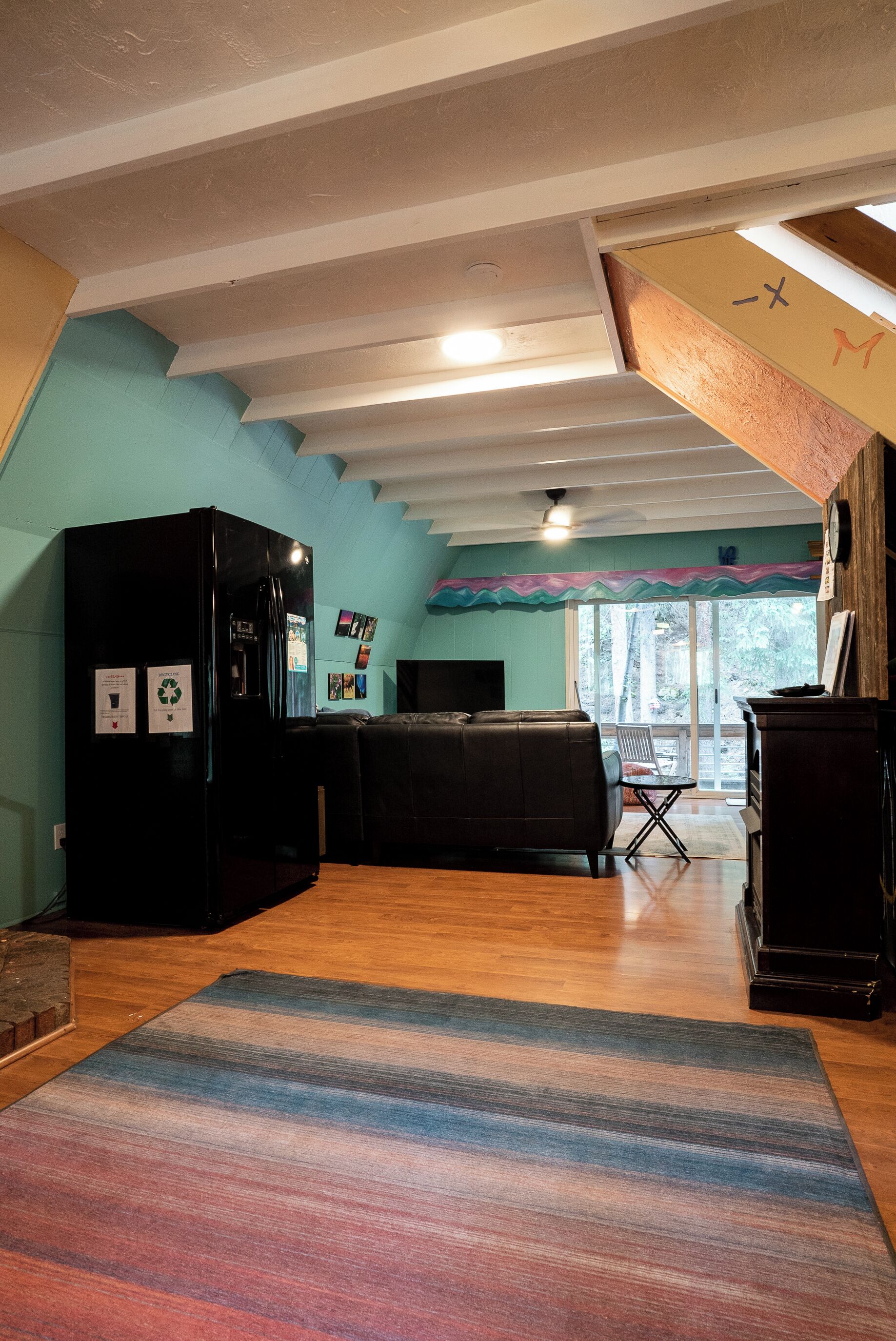 Ein Zimmer mit einem schwarzen Kühlschrank, einer Couch, einem Holzboden und einem Fenster mit Blick auf Bäume.