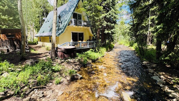 A cabin with a deck, surrounded by a forest and a stream.