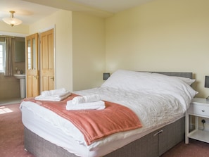 Uma cama bem arrumada com roupa de cama rosa e branca, uma mesa de cabeceira com abajur e uma porta que dá para um banheiro.