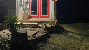 A house with a red door and white exterior walls, a small porch with steps, and a garden area with plants and a wooden bench. - Adorable one bedroom with fireplace/kitchenette (Raywick)