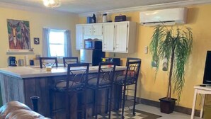 A living room with a brown leather sofa, a kitchen area with white cabinets, a dining table with chairs, and a potted plant. - Adorable one bedroom with fireplace/kitchenette (Raywick)