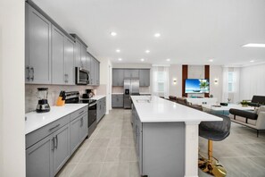 A modern kitchen with grey cabinets, a central island, and a dining area with a TV. - Seaside Paradise: 9 Bedroom Mansion 6275 (Kissimmee)