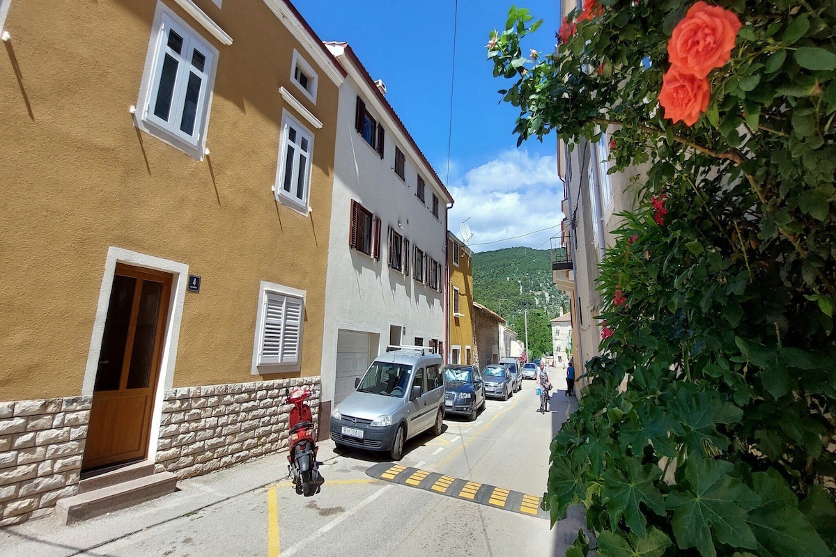 Una calle estrecha con coches aparcados y una motocicleta, edificios con balcones y un cielo despejado.