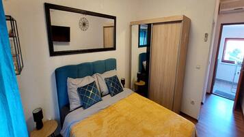 Un dormitorio con cama, mesita de noche, espejo, armario y puerta que da a otra habitación.