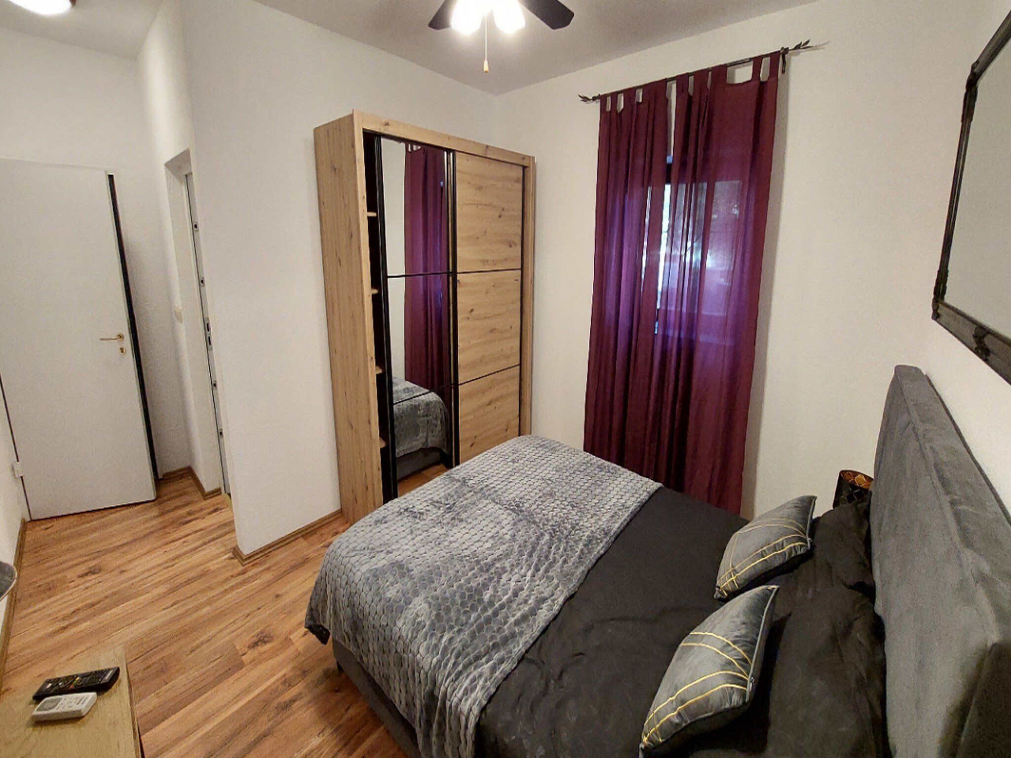 A bedroom with a bed, a wardrobe, a mirror, a ceiling fan, and a door.