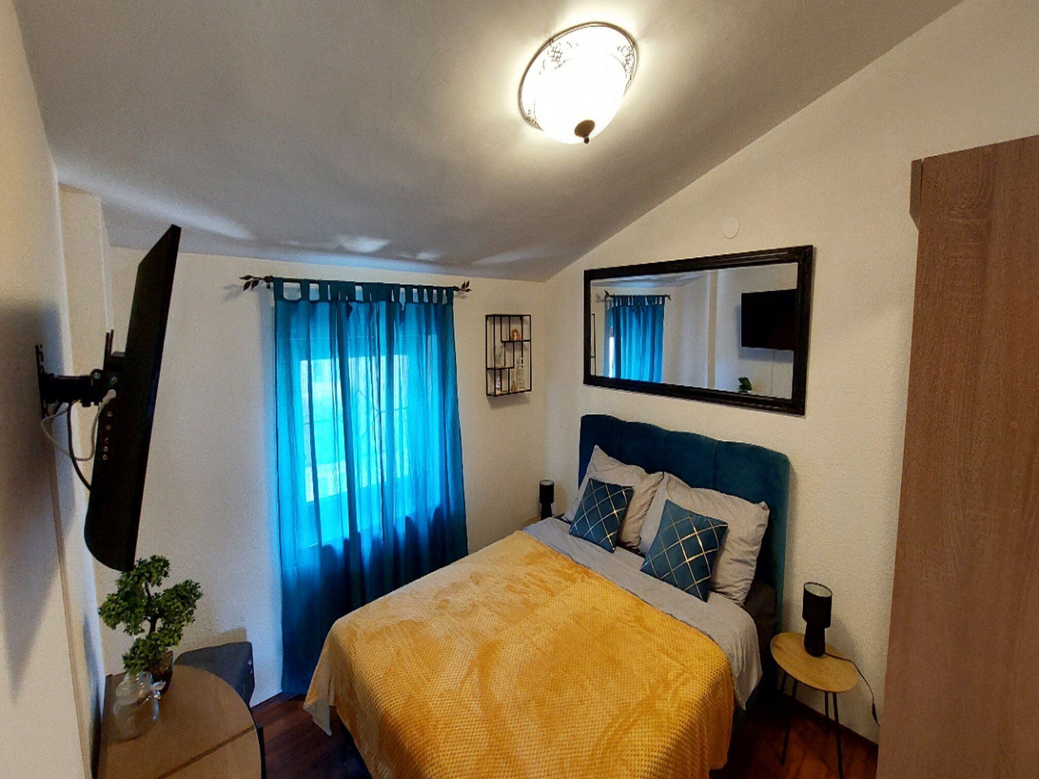 Un dormitorio con cama, mesita de noche, lámpara, espejo, ventana con cortinas y luz en el techo.
