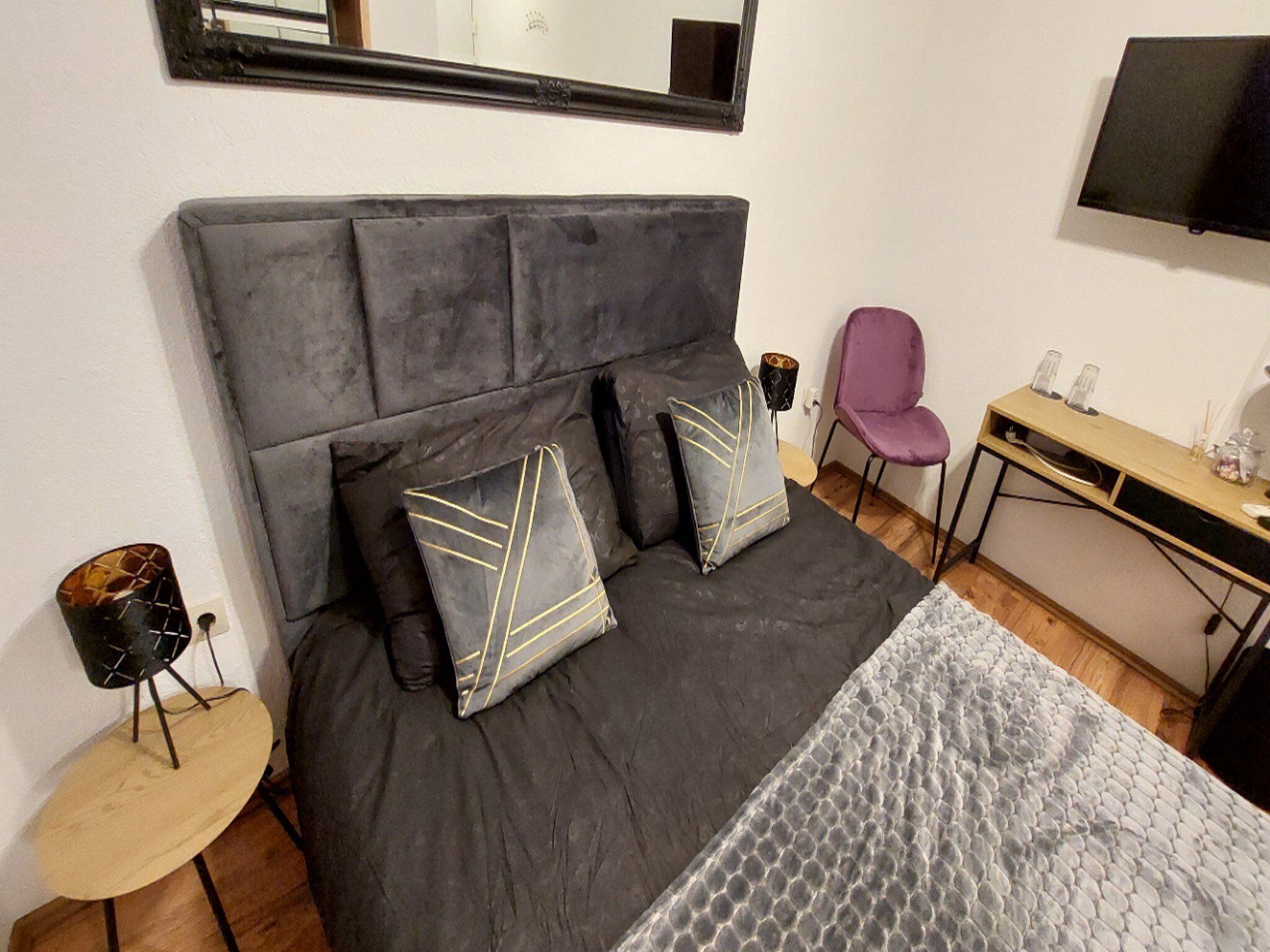 Een moderne slaapkamer met een grijs gestoffeerd bed, twee decoratieve kussens, een klein rond tafeltje, een paarse stoel, een bureau met een flatscreen-televisie en een grote spiegel aan de muur.
