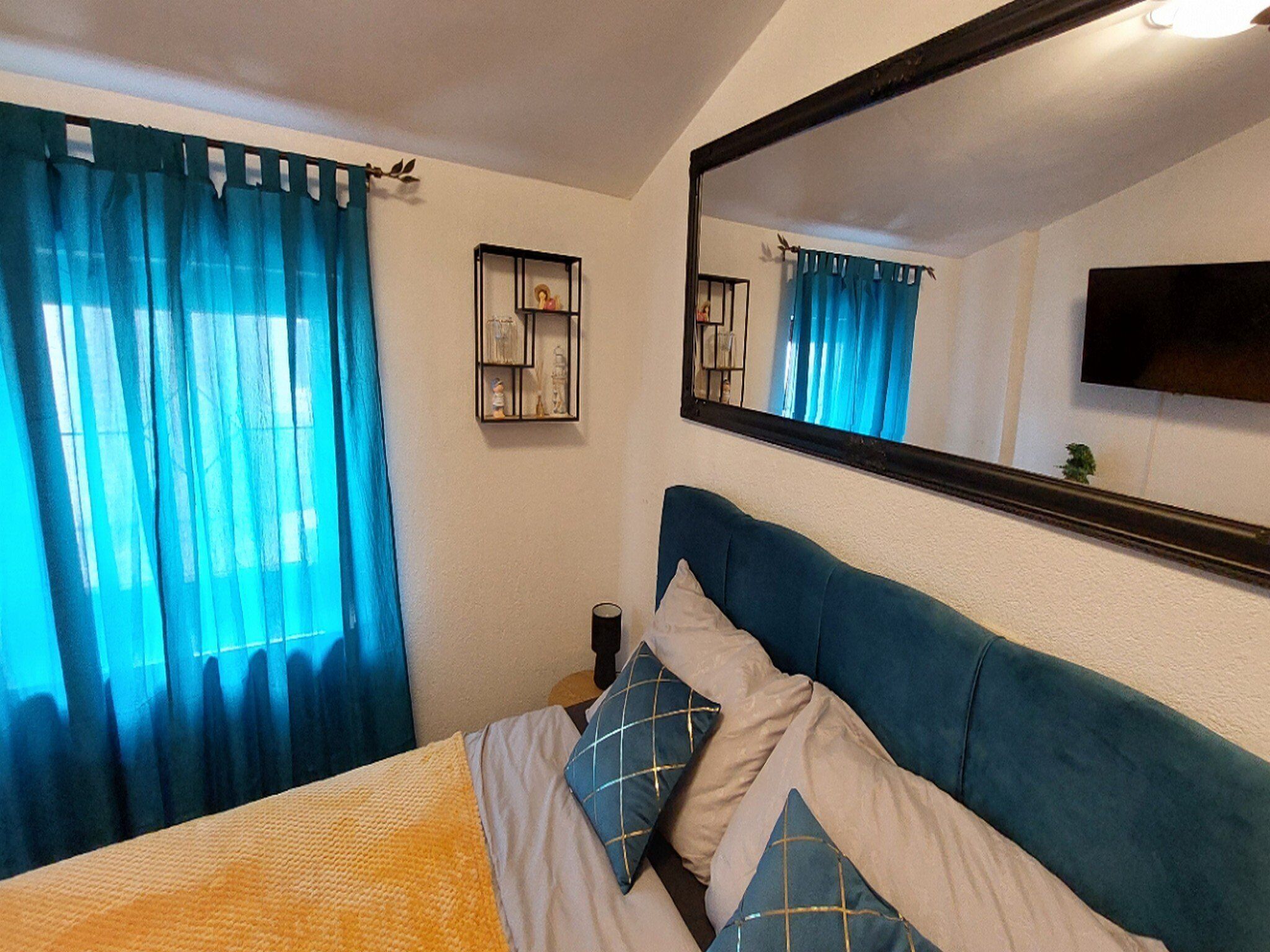 Een slaapkamer met een bed, blauwe gordijnen, een nachtkastje, een spiegel en een aan de muur bevestigde televisie.