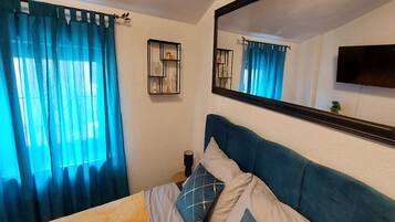 Een slaapkamer met een bed, blauwe gordijnen, een nachtkastje, een spiegel en een aan de muur bevestigde televisie.