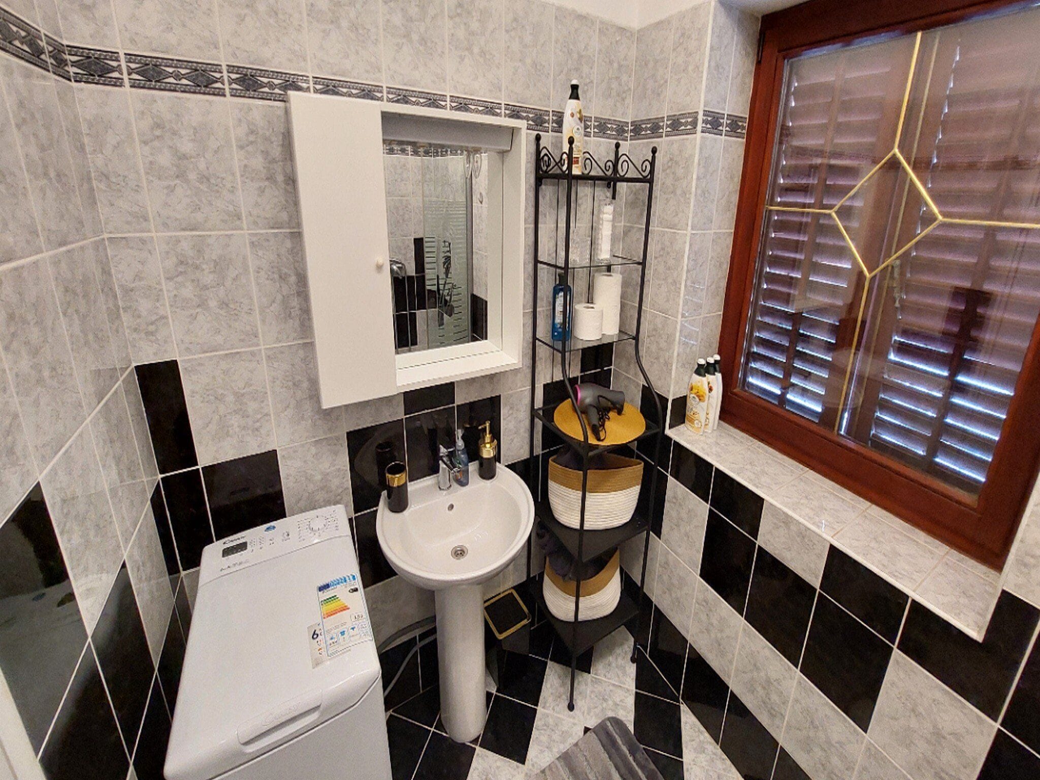 A bathroom with a white sink, a mirror, a toilet, a washing machine, and a window with blinds.