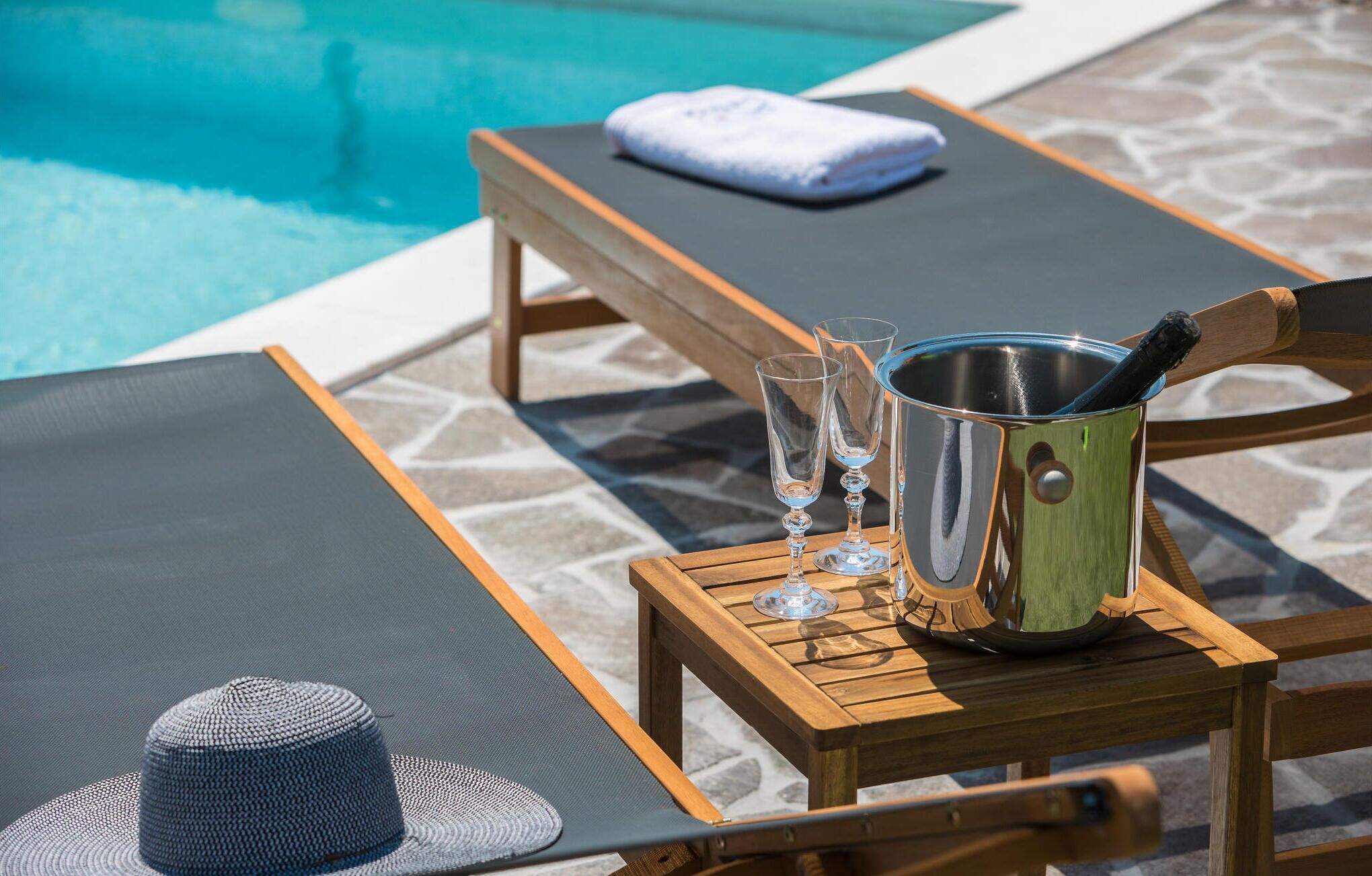 Un área de descanso junto a la piscina con una mesa de madera, un balde de metal, dos vasos y una toalla doblada.
