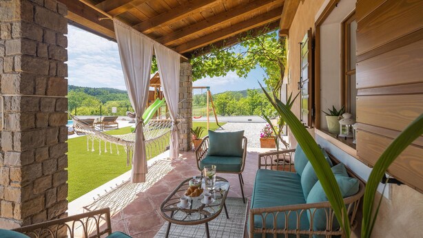 Eine überdachte Terrasse mit Korbmöbeln, einem Tisch mit einem Croissant und Blick auf einen Spielplatz und Hügel.