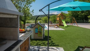 Ein Garten mit Grillplatz, einem kleinen Spielhäuschen, einer Rutsche und einer Schaukel.
