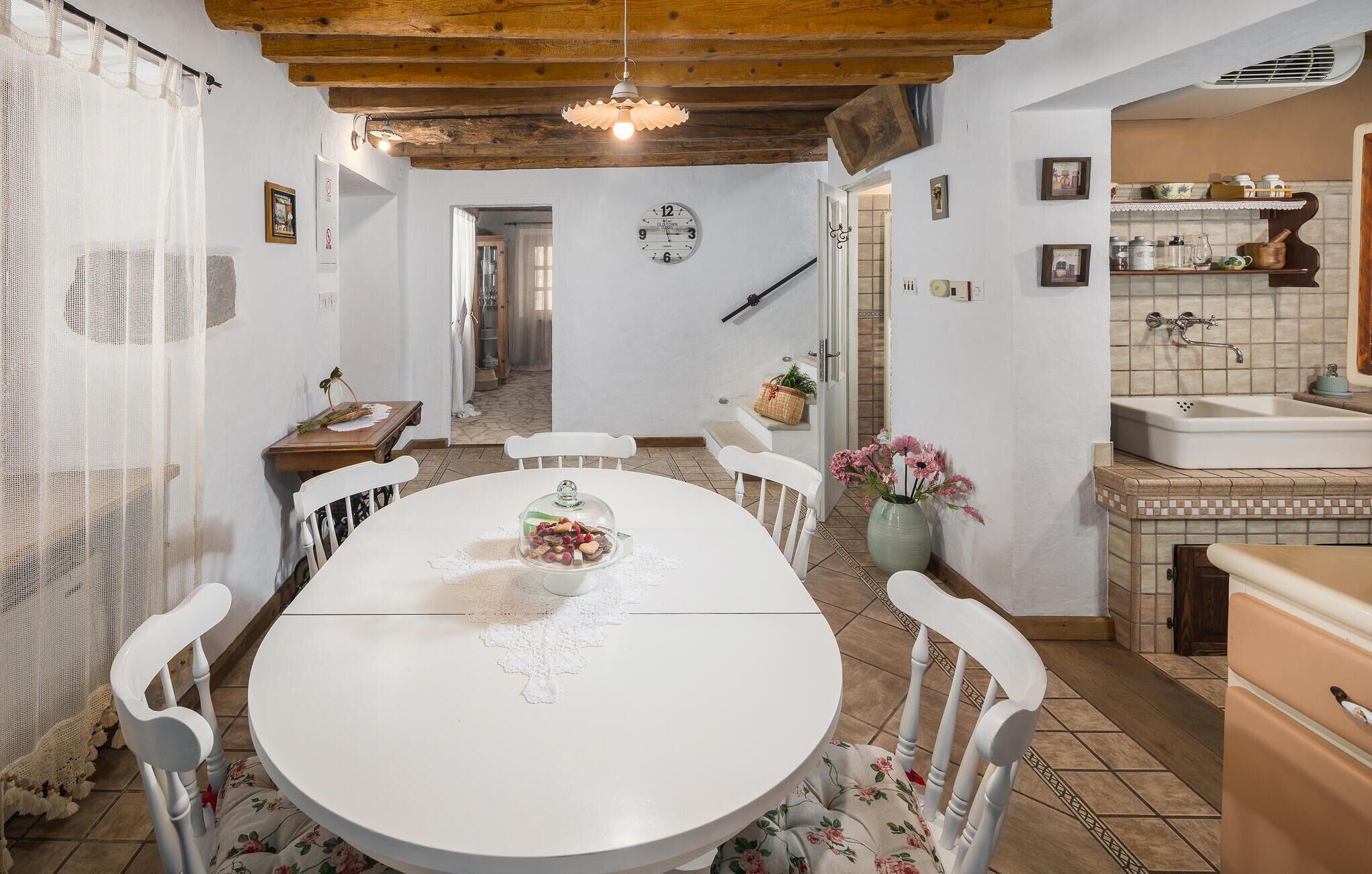 A dining area with a round table, white chairs, a chandelier, and a kitchen with a sink and shelves.