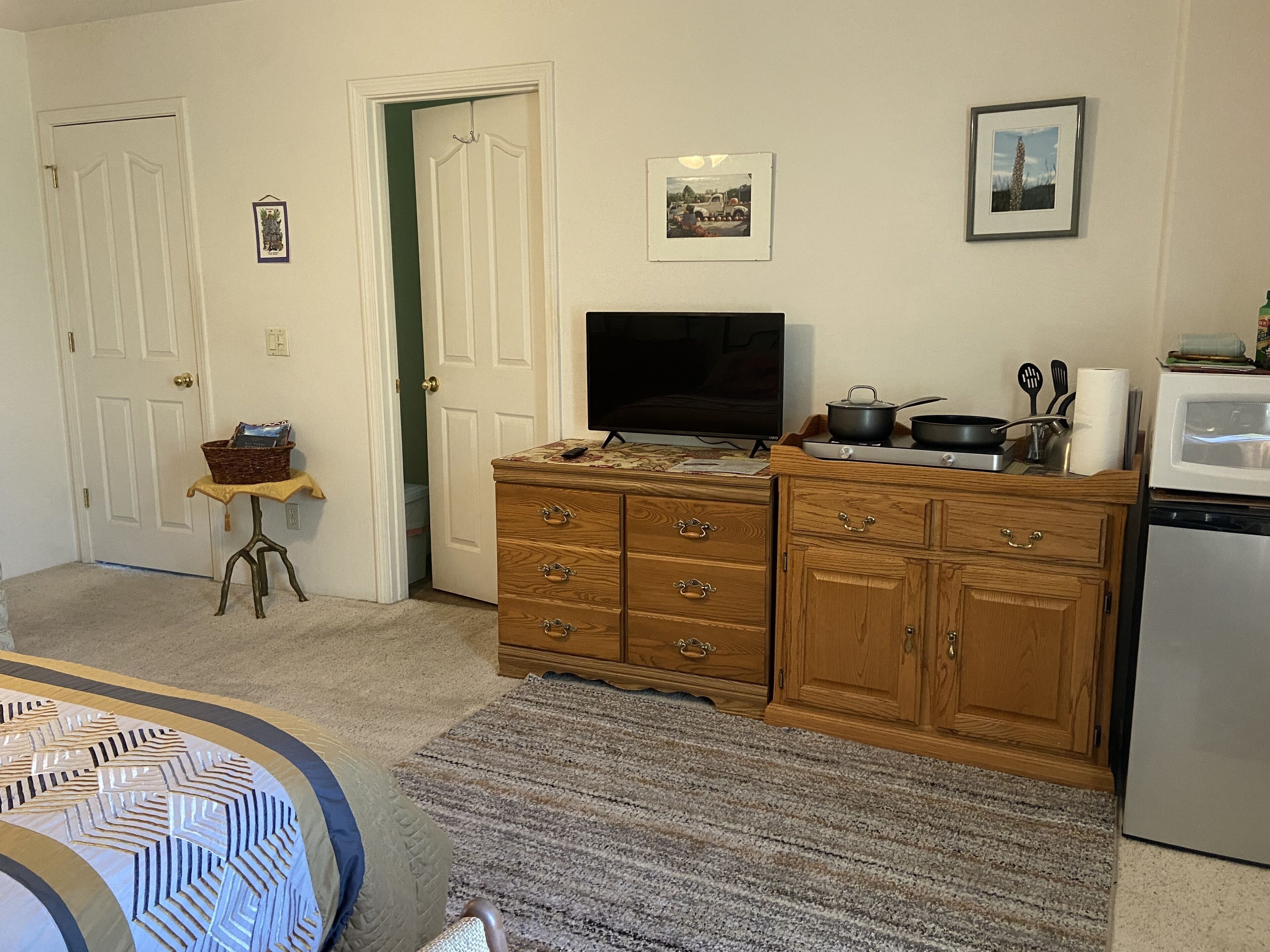 A compact kitchen with a microwave, stove, and wooden cabinets. A television is mounted above the stove, and a small table with a basket is visible near the door.