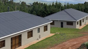 A row of modern, single-story buildings with flat roofs, surrounded by a grassy area and a dirt path. - Iter Away Highland Lodge (Nyanga)