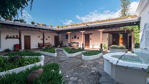 A courtyard with stone flooring, white planters with greenery, a white fountain, and a wooden bench. - Casita Tumbergia (Antigua Guatemala)
