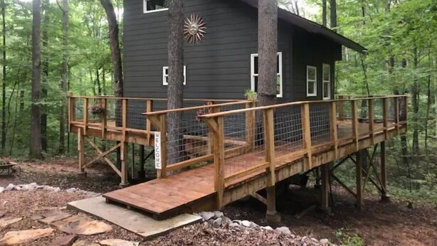 A cabin with a wooden deck and a 'WELCOME' sign, surrounded by trees.
